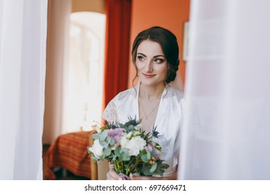 The Bride Brunette In Veil And White Satin Dressing Gown Sits By The Window