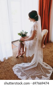 The Bride Brunette In Veil And White Satin Dressing Gown Sits By The Window