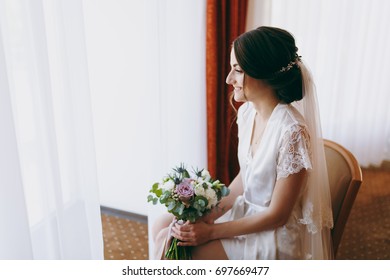 The Bride Brunette In Veil And White Satin Dressing Gown Sits By The Window