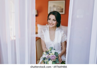 The Bride Brunette In Veil And White Satin Dressing Gown Sits By The Window