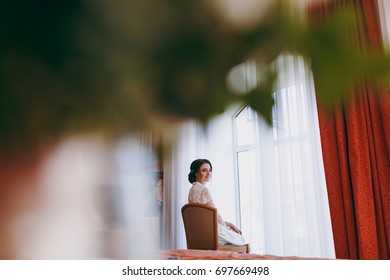 The Bride Brunette In Veil And White Satin Dressing Gown Sits By The Window