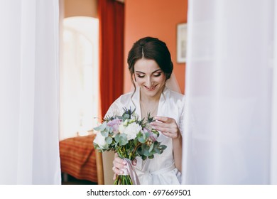 The Bride Brunette In Veil And White Satin Dressing Gown Sits By The Window