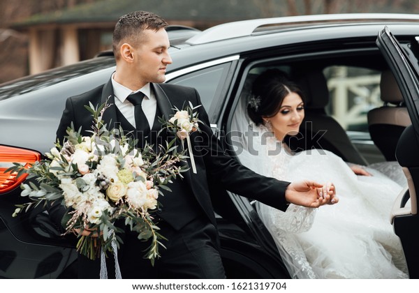 Bride with flower bouquet in car. Pretty woman\
wearing white wedding dress and tiara with veil. The bride gets out\
of the car and the groom extends a hand. Bride with wedding makeup\
and hairstyle