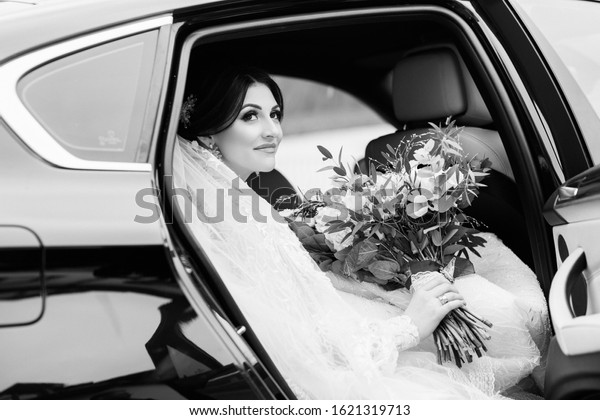 Bride with flower bouquet in car. Pretty young\
woman wearing white wedding dress and tiara with veil. The bride\
gets out of the car. Outside Photo. Beautiful bride with wedding\
makeup and hairstyle