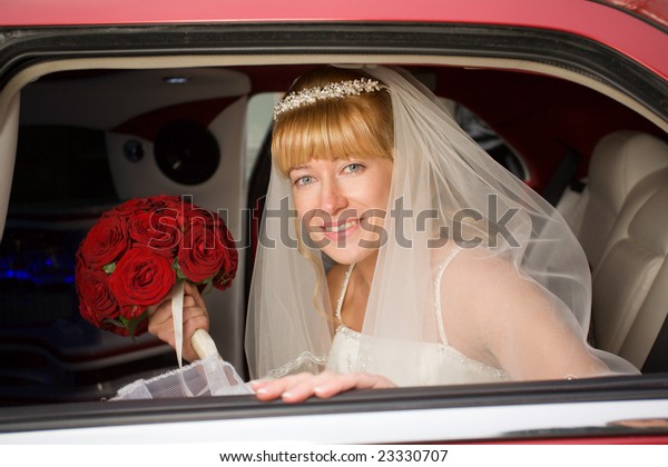 Bride with flowers inside red limo looking from\
window and smiling.
