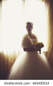 Bride In A Fur Shawl Stands Behind A Big Bright Window