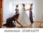 A bride is getting ready for her wedding with the help of her bridesmaids. The bride is wearing a white dress and the bridesmaids are wearing purple dresses. Scene is joyful and celebratory