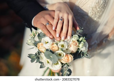 Bride and groom in black suit and wedding dress hold hands with gold wedding rings on a wedding bouquet of white and beige roses, close-up without faces