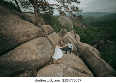 A bride and groom are climbing a rocky mountain together. The bride is wearing a white dress and the groom is wearing a blue shirt. The scene is peaceful and romantic - Powered by Shutterstock