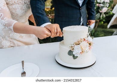 The Bride And Groom Cut A Beautiful Round Two-tiered Wedding Cake With The Word Mr And Mrs, Decorated With Flowers, Holding A Knife Together With Their Hands. Food Photography.