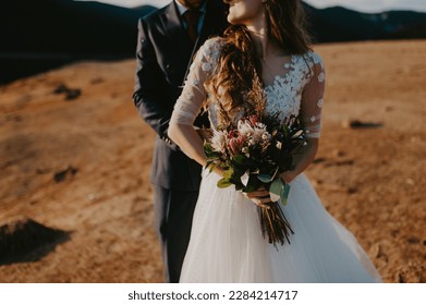 Bride and groom dressed elegant in their wedding gowns holding hands and hugging posing for their photo session in a desert like landscape by the lake. bride holding flower bouquet.