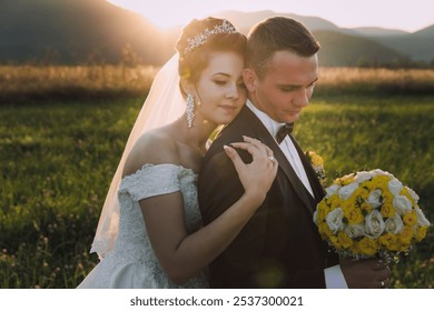 A bride and groom are embracing in a field with a bouquet of yellow flowers. The bride is wearing a veil and the groom is wearing a tuxedo. The scene is romantic and intimate, capturing the love - Powered by Shutterstock