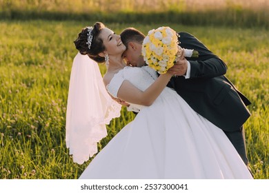 A bride and groom are embracing in a field of grass. The bride is wearing a white dress and the groom is holding a bouquet of yellow flowers. The scene is romantic and happy - Powered by Shutterstock