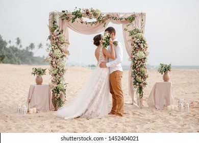 Bride and groom enjoying beach wedding in tropics, wedding arch, ocean background. Wedding ceremony on a tropical beach. Happy groom and beautiful bride kissing under the arch decorated with flowers 