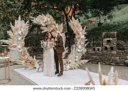 The bride and groom are enjoying themselves. Newlyweds with a wedding bouquet standing at a wedding ceremony under an arch decorated with flowers and dried flowers outdoors.