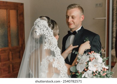 A bride and groom are getting ready for their wedding. The groom is adjusting his tie while the bride looks on. The bride is wearing a white veil and the groom is wearing a black suit - Powered by Shutterstock