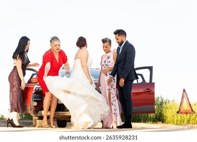 Bride, groom, and guests are fixing a problem with their car on the side of the road during a wedding