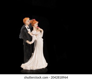 Bride And Groom Holding An Old Cake Topper On A Black Background. Figurines For A Wedding Cake. Figures Of Newlyweds In Festive Costumes.