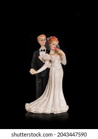 Bride And Groom Holding An Old Cake Topper On A Black Background. Figurines For A Wedding Cake. Figures Of Newlyweds In Festive Costumes.