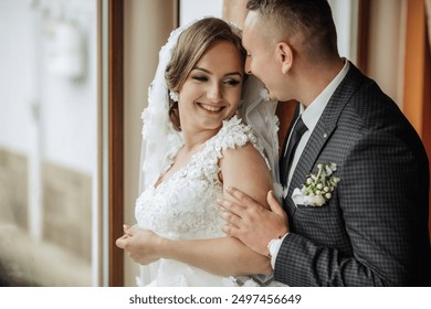 A bride and groom are hugging each other in a window. The bride is wearing a white dress and the groom is wearing a suit. Scene is happy and loving - Powered by Shutterstock