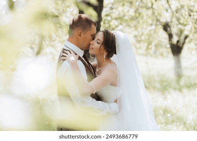 A bride and groom are hugging in a field. The bride is wearing a white veil and the groom is wearing a red vest - Powered by Shutterstock