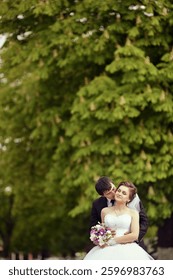 A bride and groom are hugging in front of a tree. The bride is wearing a white dress and the groom is wearing a suit