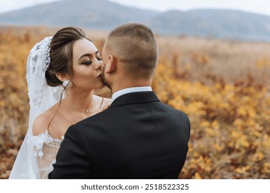 A bride and groom kiss in a field. The bride is wearing a veil and the groom is wearing a suit. Scene is romantic and happy - Powered by Shutterstock