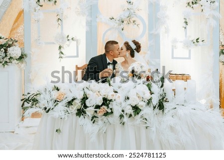 A bride and groom are kissing at a wedding reception. The bride is wearing a white dress and the groom is wearing a black suit. The table in front of them is covered with flowers