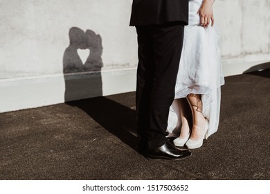 Bride And Groom On Their Wedding Forming A Heart With The Shadow Of Their Faces On A Wall. In Front, You See The Feet Of The Bride. She Is Lifting Her Dress