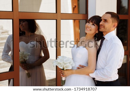 The bride and groom on the wedding day are standing near the restaurant, the groom hugs the bride from behind. The bride holds a beautiful bouquet. Reflection of the bride and groom in the window.