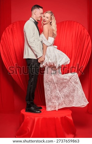Bride and groom posing before red heart backdrop. Luxury wedding fashion, romantic relationship mood and clean copy space for invitation or engagement promotion.