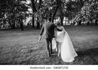 Bride And Groom Running On Green Grass Of Golf Course, Back View. Happy Wedding Couple Walking In The Forest. Black And White Photo