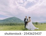 A bride and groom stand in a field with a mountain in the background. The bride is wearing a white dress and the groom is wearing a suit