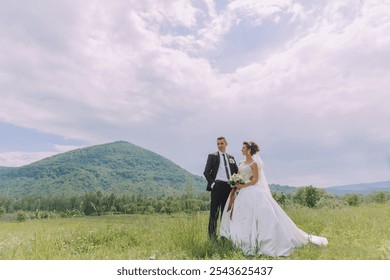 A bride and groom stand in a field with a mountain in the background. The bride is wearing a white dress and the groom is wearing a suit - Powered by Shutterstock