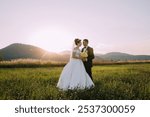 A bride and groom stand in a field with mountains in the background. The bride is wearing a white dress and the groom is wearing a black suit. They are holding hands and smiling at the camera