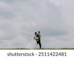 A bride and groom are standing in a field on a cloudy day. The bride is wearing a white dress and the groom is wearing a suit. They are holding hands and looking at the camera