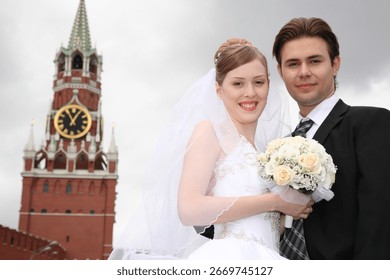 A bride and groom are standing in front of a clock tower, with the bride holding a bouquet of flowers. Scene is romantic and happy, as the couple is celebrating their wedding