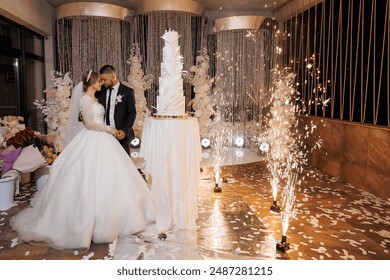 A bride and groom are standing in front of a wedding cake. The bride is wearing a white dress and the groom is wearing a suit. Scene is happy and celebratory - Powered by Shutterstock