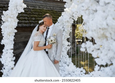 A bride and groom are standing in front of a white archway, with the bride holding a bouquet. The scene is a wedding ceremony, and the couple is dressed in formal attire. Scene is joyful and romantic - Powered by Shutterstock