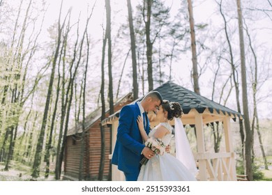 A bride and groom are standing in front of a wooden gazebo, with the bride holding a bouquet - Powered by Shutterstock