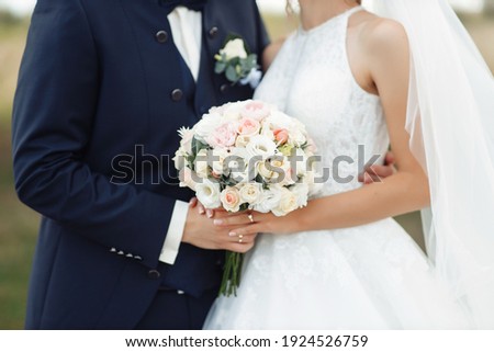 The bride and groom are standing next to each other and a man in a blue stylish suit with a bow-tie gently holds her by the shoulders. Girl in a white dress with a bouquet of roses flowers in her hand