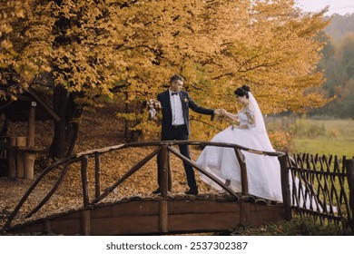 A bride and groom are standing on a bridge, holding hands and looking at the camera. The scene is set in a beautiful autumn setting, with leaves falling around them. Scene is romantic and intimate - Powered by Shutterstock