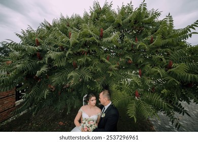 A bride and groom are standing under a large tree, with the bride holding a bouquet. Scene is romantic and intimate, as the couple shares a special moment together - Powered by Shutterstock