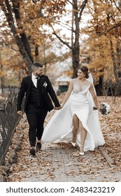 A bride and groom are walking down a path in the fall. The bride is wearing a white dress and the groom is wearing a suit. They are holding hands and appear to be happy