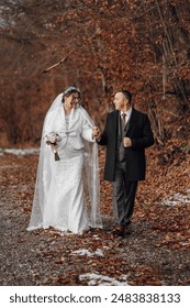A bride and groom are walking down a path in the woods. The bride is wearing a white dress and a fur stole, while the groom is wearing a black suit. They are holding hands and smiling
