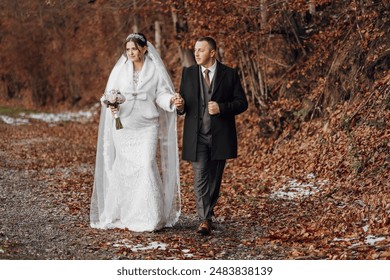 A bride and groom are walking down a path in the woods. The bride is wearing a white dress and a fur stole, while the groom is wearing a suit. They are holding hands