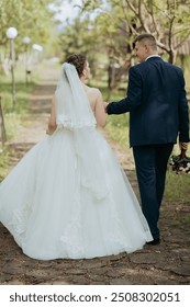 A bride and groom are walking down a path in a garden. The bride is wearing a white dress and a veil, while the groom is wearing a suit. They are holding hands and appear to be happy and in love
