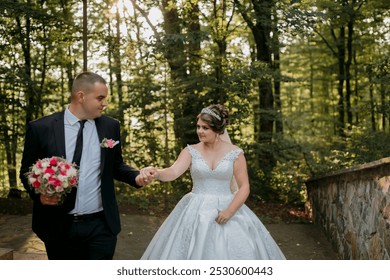A bride and groom are walking down a path in a forest. The bride is wearing a white dress and the groom is wearing a suit. The bride is holding a bouquet and the groom is holding her hand