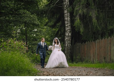 A bride and groom are walking down a path in the woods. The bride is wearing a white dress and the groom is wearing a suit. They are holding hands and appear to be enjoying their walk together