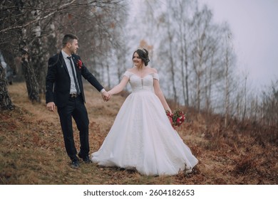A bride and groom are walking down a path in the woods. The bride is wearing a white dress and the groom is wearing a suit. The couple is holding hands and the bride is holding a bouquet